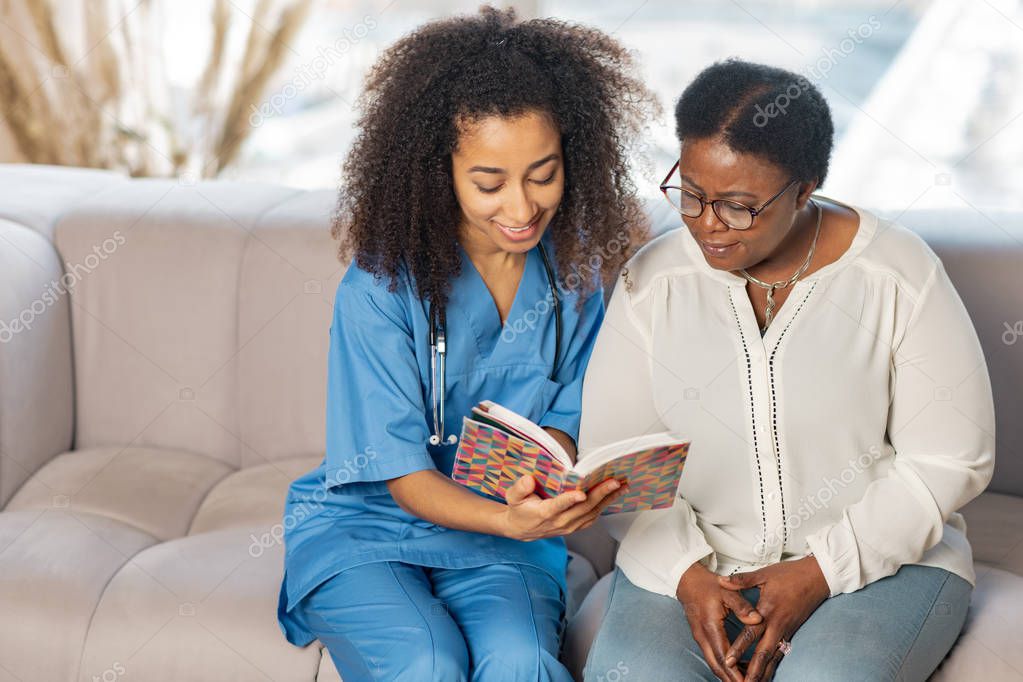 Caring nurse sitting on sofa near patient and reading book for her