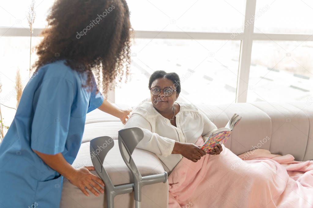 Aged woman having rest on sofa and looking at caring nurse