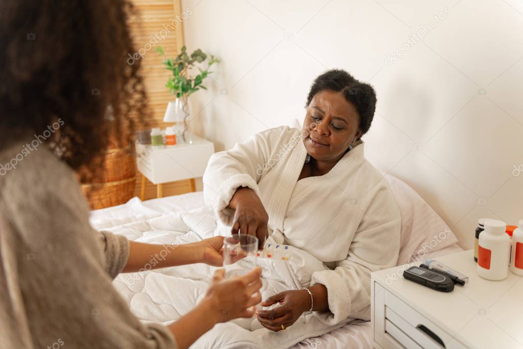 Woman lying in bed taking pills and water from caring nurse