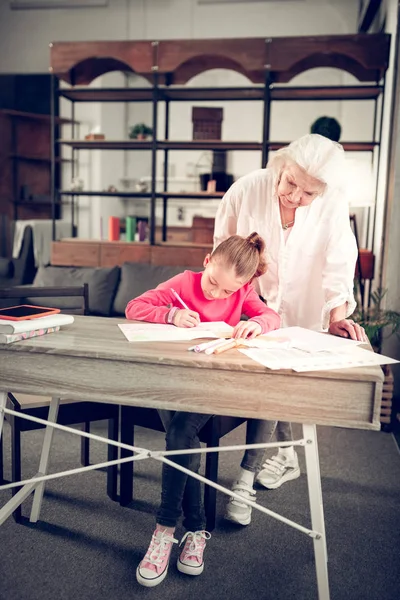 Granny watching her girl sitting at the table and doing homework — Stock Photo, Image