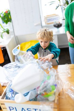 Blonde-haired boy standing near box with plastic after sorting waste clipart