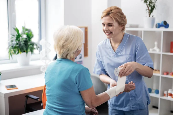 Χαρούμενη θετική νοσοκόμα τυλίγοντας επίδεσμο γύρω από τους ασθενείς της χέρι — Φωτογραφία Αρχείου