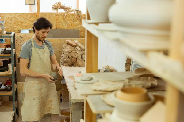 Concentrated dark-haired guy in long apron rolling clay — Stock Photo, Image