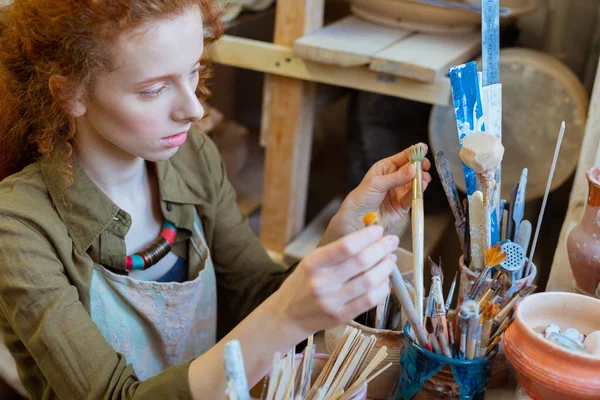 Serious curly woman sorting out paintbrushes in big pile — Stock Photo, Image