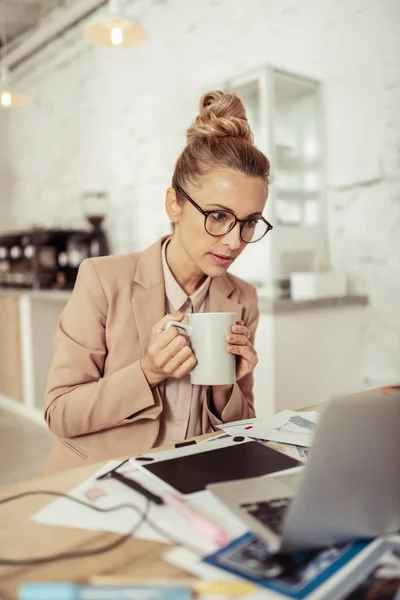 Mulher concentrada bebendo café e olhando para seu laptop . — Fotografia de Stock