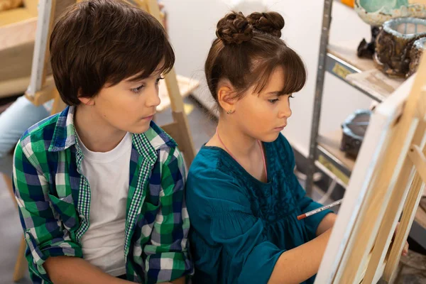 Irmão de cabelos escuros assistindo sua irmã pintando na escola de arte — Fotografia de Stock