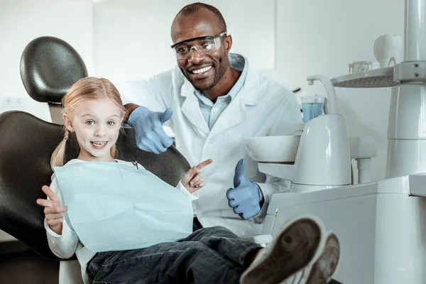 Dentista afro-americano de uniforme completo sentado atrás do seu pequeno paciente — Fotografia de Stock