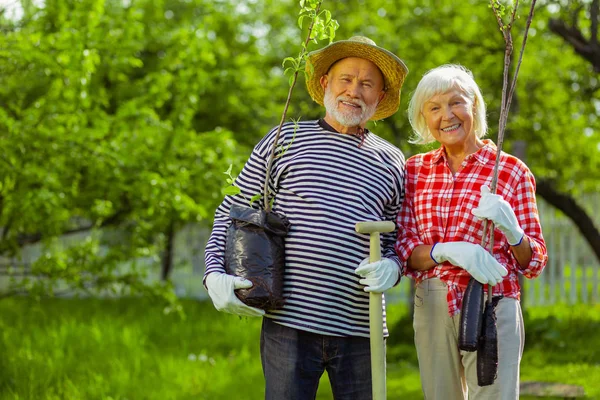 Retired husband and wife holding trees and shovel standing in the garden