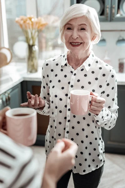 Mujer mayor de moda sonriendo y luciendo feliz mientras bebe té —  Fotos de Stock