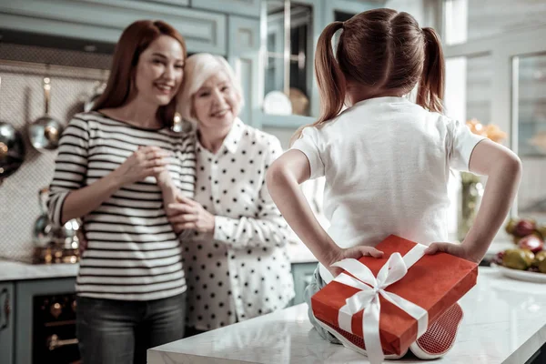 Милая девушка сидит на столе и прячет подарок от мамы и бабушки — стоковое фото