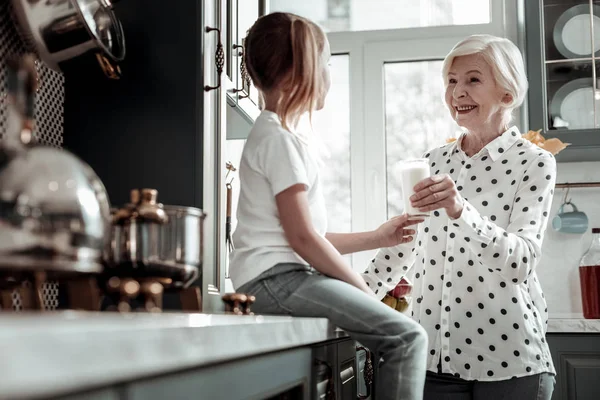 Liebende Großmutter, die in der Küche steht und dem Mädchen Milch anbietet — Stockfoto