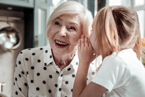 Cute little girl leaning to her happy granny and whispering secret information — Stock Photo, Image