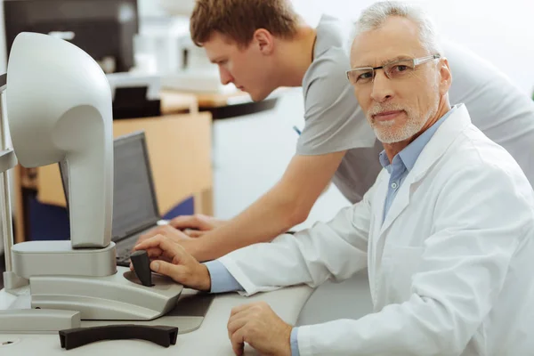 Grey-haired bearded ophthalmologist sitting near his assistant — Stock Photo, Image