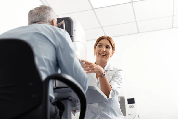 Beautiful young eye doctor smiling while examining retired man — Stock Photo, Image