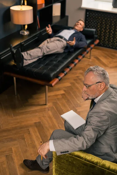Задумчивый серьезный психотерапевт сидит рядом со своим пациентом — стоковое фото