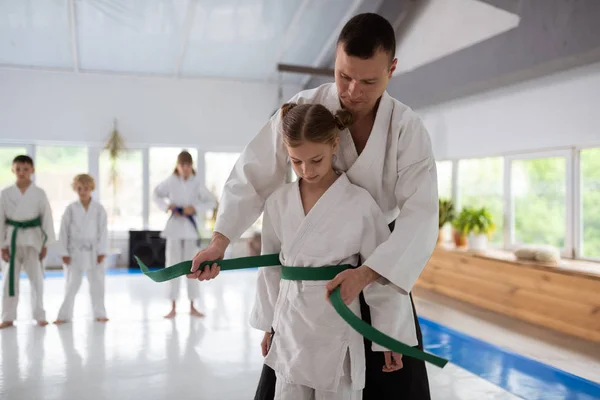 Dark-haired aikido trainer putting belt on waist of girl — Stock Photo, Image