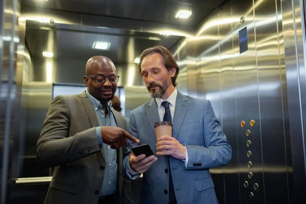 Dos hombres de negocios que leen información en el teléfono inteligente mientras usan el ascensor — Foto de Stock