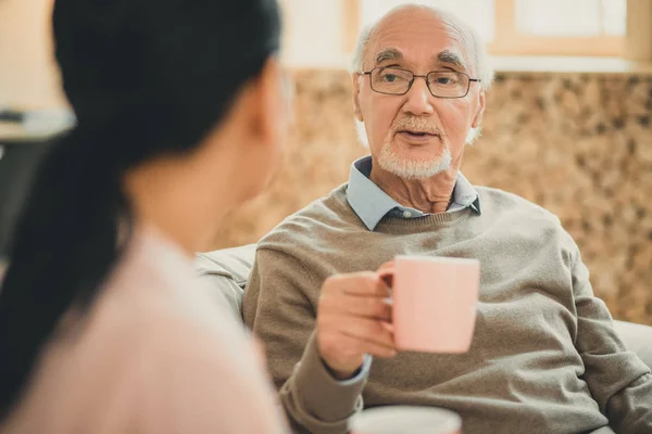 Wise old man with pink cup speaking to brown-haired lady