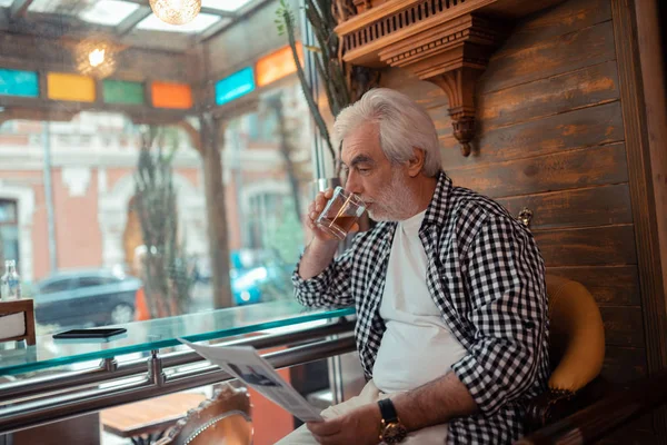 Uomo dai capelli grigi che beve whisky e legge notizie — Foto Stock