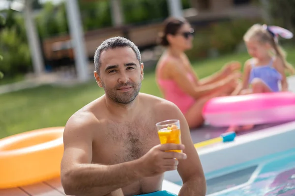 Hombre bebiendo cóctel frío mientras se enfría con la familia cerca de la piscina — Foto de Stock