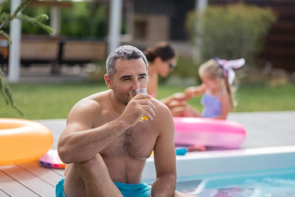 Hombre de pelo gris bebiendo cóctel sentado cerca de la piscina con la familia — Foto de Stock