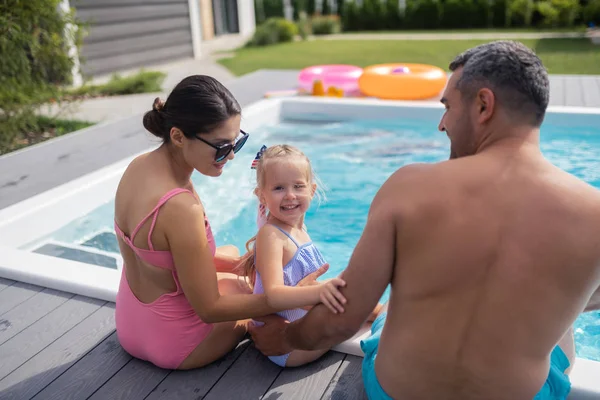 Hija sonriendo mientras descansa cerca de la piscina con mamá y papá — Foto de Stock