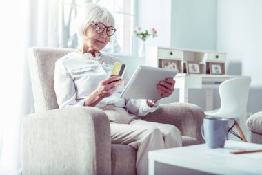 Tablet üzerinden online alışveriş yaparken banka kartı tutan modern emekli kadın