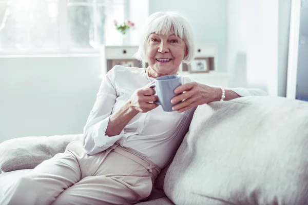 Smilende, eldre dame med fin, hvit bluse som sitter i stua – stockfoto