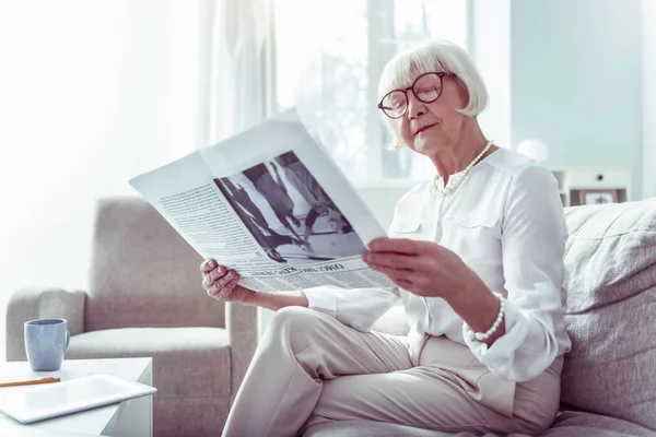 Krásná starší dáma na brýlích, čteš ranní noviny na pohovce — Stock fotografie