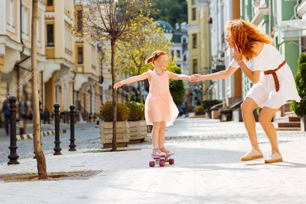 Be careful. Joyful red haired woman wearing white dress while walking down the street with her child