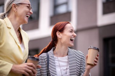 Birlikte mutlu. Bel-up kağıt bardak ile iki memnun iş bayanlar açık havada gülüyor ve mesafe içine bakarak