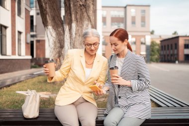 Bankta. Genç kızıl saçlı kadın dışarıda bankta otururken gri saçlı meslektaşına resim gösteren