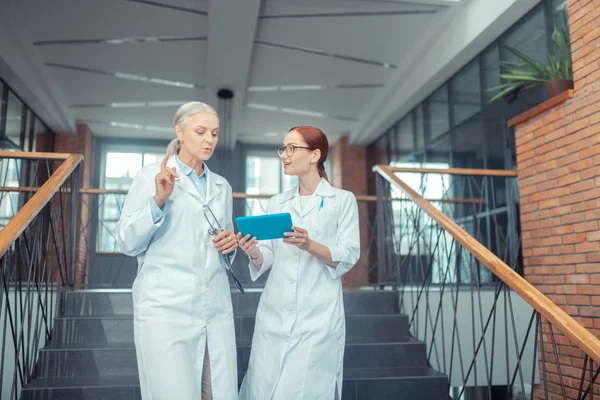 Female doctors. Senior Caucasian female doctor standing on the stairs in a modern medical establishment while talking to her young colleague