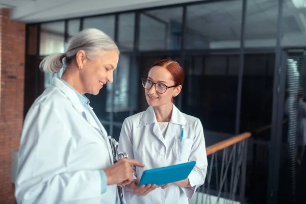 两个医生一位面带微笑的高级女医生侧视 她的同事旁边走廊上站着一块药片 — 图库照片