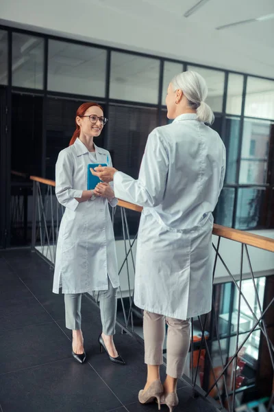 女医生一位白发苍苍的女医生站在走廊里 站在她年轻的微笑的同事面前的后视图 — 图库照片