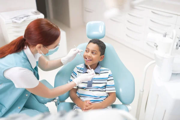 Niño de ojos oscuros abriendo la boca mientras el dentista examina la cavidad —  Fotos de Stock