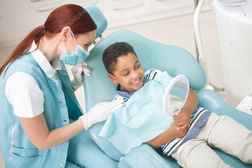 Boy looking at teeth into mirror while sitting in dental chair