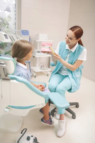Дівчина слухає стоматолога, показуючи їй, як користуватися зубною щіткою — стокове фото