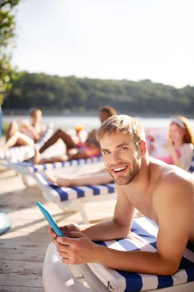 Man reading online news and sunbathing near river with friends