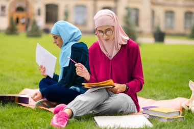 Çimenlerin üzerinde oturan ve materyali tekrarlayan iki Müslüman öğrenci