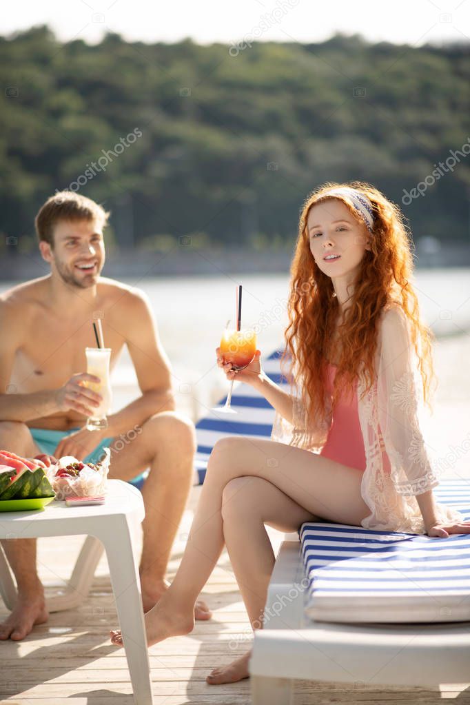 Red-haired girlfriend holding glass of cocktail sitting near her man