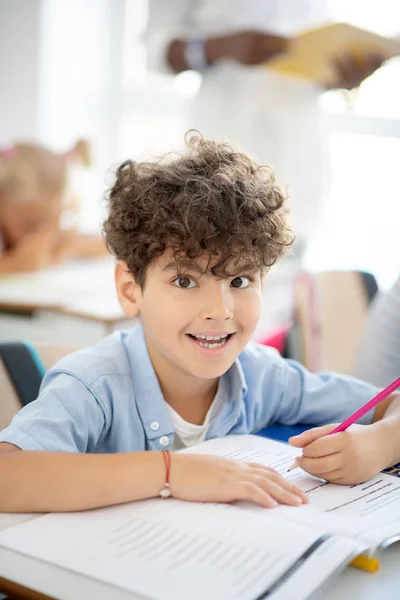 Dunkeläugiger Schuljunge macht beim Schreiben lustiges Gesicht — Stockfoto