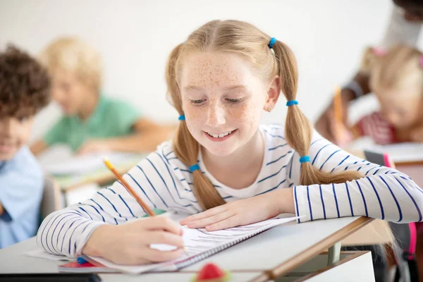 Glimlachend meisje met sproeten glimlachend tijdens het schrijven op de les — Stockfoto