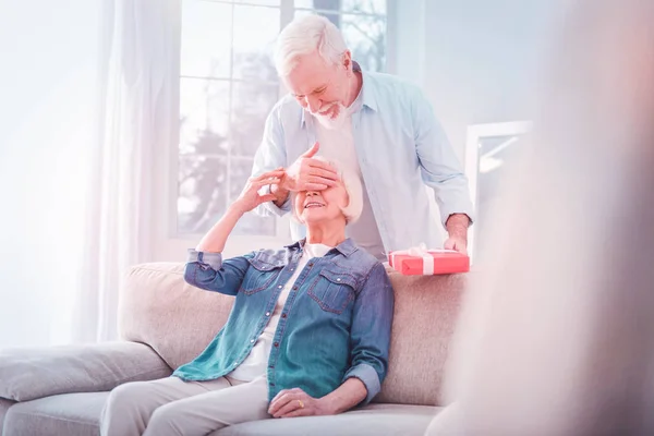 Bärtiger Mann kommt zu seiner Frau auf Sofa sitzend und präsentiert Überraschung — Stockfoto