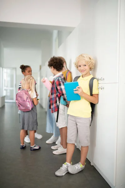 Niño con la tableta azul de pie cerca de amigos en el pasillo de la escuela — Foto de Stock