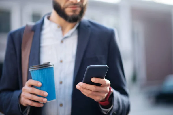 Empresario barbudo leyendo e-mail en el teléfono y sosteniendo la taza de café —  Fotos de Stock