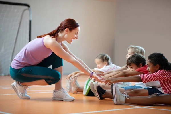 PE profesor usando leggings sonriendo mientras ayudar a chica estiramiento — Foto de Stock