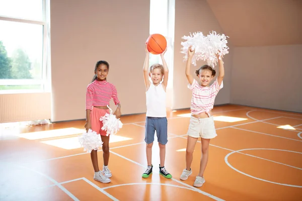 Petites pom-pom girls mignonnes debout près de camarade de classe avec balle — Photo