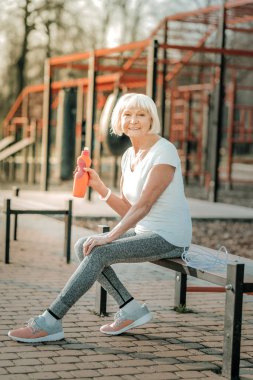 Spor sahasında. Enerjik gülümseyen yaşlı bayan spor salonu giyim giyen , sağ elinde su şişesi tutan ve spor bankında oturan