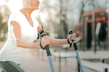 Yürüyüş direkleri ile egzersiz. Spor alanında yürüyüş direkleri ile çalışan ve kulaklıkile müzik dinleyen kısa gri saçlı güçlü yaşlı sportif bayan